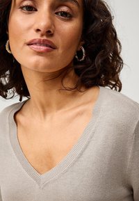 Maglione beige chiaro con scollo a V, collare a coste e tessuto morbido, abbinato a orecchini a cerchio in tonalità oro.