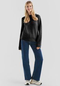 Uśmiechnięta kobieta w oversize'owym czarnym swetrze, niebieskich dżinsach, białych sneakersach i okularach, stojąca na tle gładkiego jasnoszarego tła.