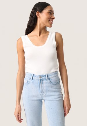 Mujer sonriente con cabello largo y oscuro lleva una camiseta blanca sin mangas y vaqueros azules claros de talle alto con pequeños adornos.