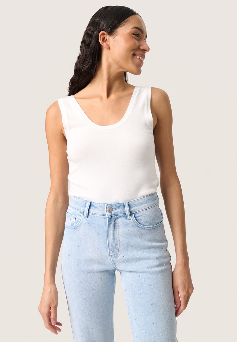 Femme souriante aux longs cheveux foncés porte un haut blanc sans manches et un jean taille haute bleu clair avec de petites décorations.