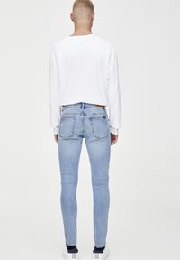 Jeans ajustados de color azul claro con una textura de denim suave, dos bolsillos traseros y un parche de cuero marrón en la cintura, combinados con una camisa blanca de manga larga.