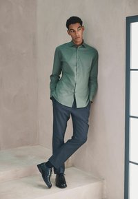 Hellgrünes Hemd mit Knöpfen und engem Kragen, kombiniert mit dunkelgrauer, maßgeschneiderter Hose und schwarzen Lederschuhen, vor einem neutralen Hintergrund stehend.