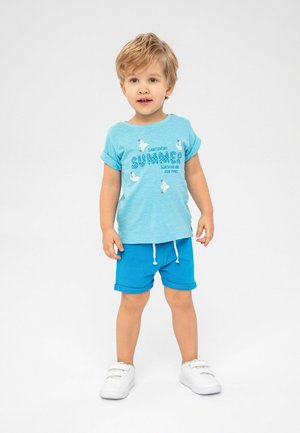 Hellblaue Kurzarm-T-Shirt mit weißen grafischen Text, kombiniert mit blauen Shorts. Das Outfit wird durch weiße Schuhe mit Klettverschlüssen ergänzt.