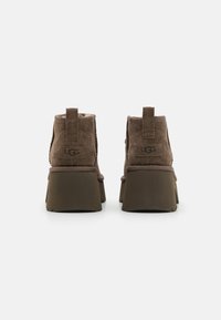 Botas de tobillo de ante marrón con una suela y tacón de plataforma gruesos y oscuros. Presentan una etiqueta en la parte posterior y el logo de UGG en el lateral.