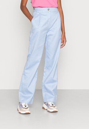 Pantalones de tiro alto en azul claro, con piernas rectas, pliegues y cierre de botón en el frente, combinados con zapatillas blancas y marrones.