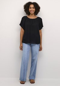 Smilende kvinde med krøllet hår iført en løs sort bluse, lyseblå, vide jeans og brune sandaler, stående mod en ensfarvet væg.