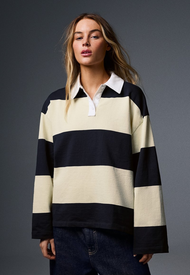 Femme aux longs cheveux portant un maillot de rugby ample rayé noir et beige avec col blanc et jean bleu foncé sur fond gris.
