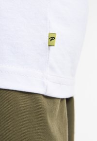 Tricou alb cu o etichetă mică verde care are un "P." negru. Marginile tricoului sunt simple, purtat cu pantaloni texturati de culoare verde măslinie.