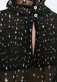 Sort gennemsigtig top med hvide prikker, der har en rynket tekstur, rund halsudskæring og en knappelukning foran med en nøglehuldetalje.