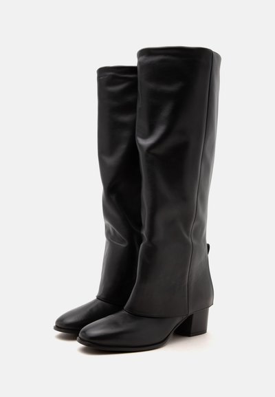 Bottes en cuir noir hautes jusqu’au genou, avec un bout arrondi et un talon bloc, placées côte à côte sur un fond blanc uni.