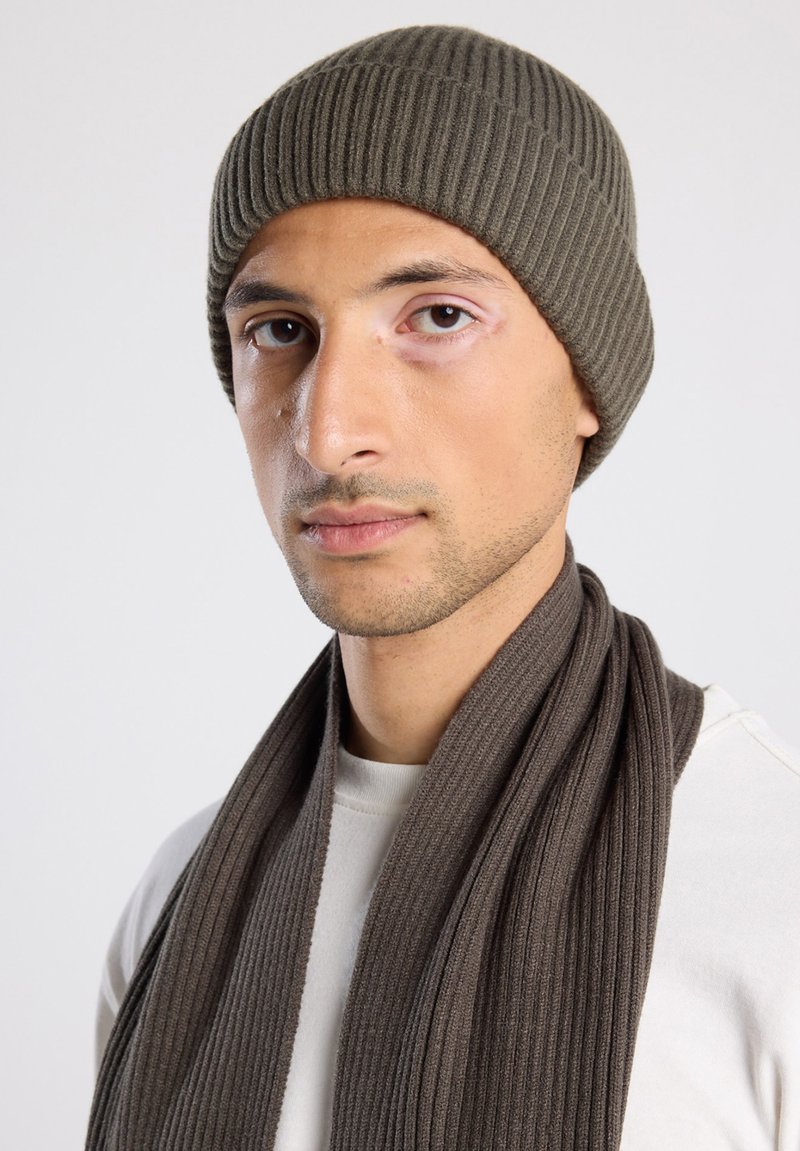 Brown ribbed knit beanie and matching ribbed scarf, both made of soft material, worn over a light-colored top. Textured finish and snug fit.