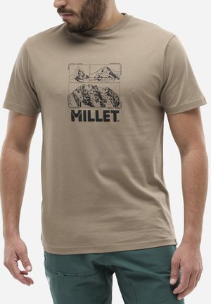 Homme portant un t-shirt beige avec des illustrations graphiques de montagnes noires et le mot "MILLET" sur le devant, porté avec un pantalon vert.