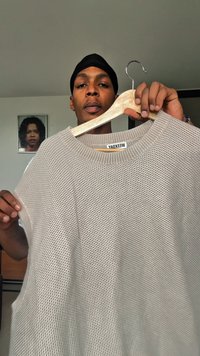 Bărbat îmbrăcat cu un batic negru ține un pulover din tricot bej texturat pe un umeraș din lemn într-un mediu interior, cu un portret înrămat în fundal.