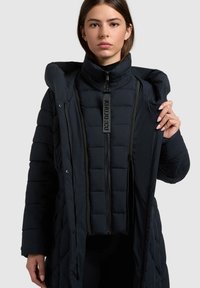 Jonge vrouw die gelaagde zwarte gewatteerde jassen draagt, en de buitenste jas opent om de binnenste jas met grote rits aan de voorkant te laten zien.