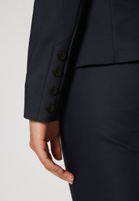 Gros plan sur la main d'une personne reposant à côté de la manche d'un blazer sombre avec quatre boutons noirs sur le poignet.