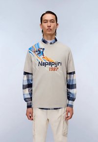 Beige katoenen T-shirt met blauw en oranje grafisch ontwerp, met de tekst "Napapijri" en "1987." Gelaagd met een geruit blauw-wit overhemd.