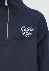 Navy blue sweatshirt with high collar, zip closure, and embroidered "Calvin Klein" logo in light blue. Smooth texture, fitted design.