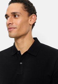 Jonge man met kort gekruld haar, gekleed in een zwart poloshirt met kraag, kijkt naar links met een lichte glimlach tegen een witte achtergrond.