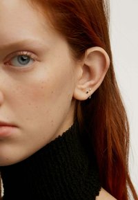 Pendientes de oro con una gema azul y un pequeño acento de estrella, exhibidos en la oreja de una persona. La persona tiene el cabello rojo y un cuello de tortuga negro texturizado.