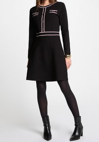 Abito nero con maniche lunghe, caratterizzato da dettagli rosa sul colletto e tasche, un corpetto aderente e una gonna svasata. Stivaletti neri a caviglia.
