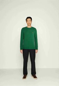 Vīrietis stāv pretī vienkāršai fona, ģērbies zaļā džemperī, melnās bikses un brūnās kedās, skatoties uz priekšu ar neitrālu izteiksmi.