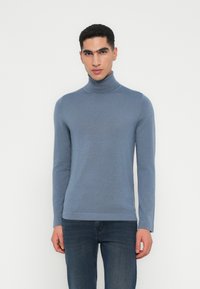 Junger Mann in einem taillierten blauen Rollkragenpullover und dunkelbblauen Jeans, der vor einem schlichten weißen Hintergrund steht.