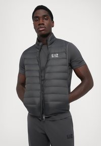 Gilet rembourré gris avec un col haut, fermeture éclair, et logo EA7 sur la poitrine gauche, porté sur une chemise à manches courtes assortie.