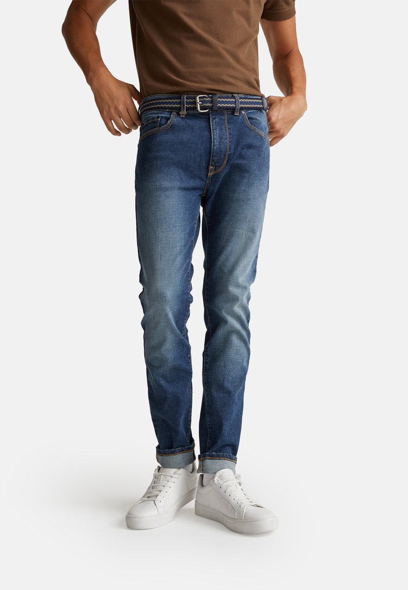 Jeans in denim blu con una finitura sbiadita, caratterizzati da una cintura intrecciata e orli arrotolati, abbinati a sneakers bianche su uno sfondo neutro.