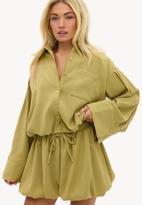 Femme aux longs cheveux blonds portant une chemise boutonnée oversize vert olive et un short taille haute assorti avec cordon, posant avec la main sur la hanche.