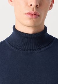 Tamno plavi rolak pulover od mekog, rebrastog materijala. Ima uski ovratnik i glatku teksturu bez vidljivih uzoraka.