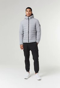 Grijze gewatteerde jas met ritssluiting en capuchon, met verticale stiknaden en twee voorzakken, gecombineerd met zwarte cargobroek en witte sneakers.