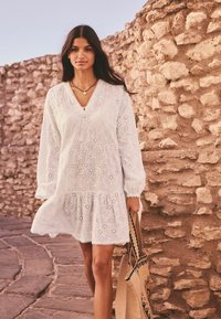 Robe blanche en dentelle avec un décolleté en V, des manches longues et un ourlet froncé. Associée à un sac fourre-tout à motifs. Fond en mur de pierre.