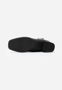 Zapato de cuero negro con punta cuadrada, tacón bajo y textura suave, con un botón decorativo en la parte superior. Suela de goma plana.