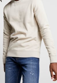 Sweatshirt beige à manches longues, col et poignets côtelés, doté d'une texture lisse. Associé à un jean bleu en denim, présentant une légère usure.