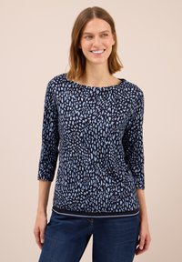 Frau trägt ein navyblaues Oberteil mit hellblauen unregelmäßigen Punkten und Dreiviertelärmeln, kombiniert mit dunkelblauen Jeans, lächelt entspannt mit relaxed Händen.