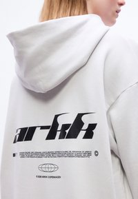 Άτομο που φοράει λευκό φούτερ με κουκούλα με μεγάλο μαύρο λογότυπο "arkk" και μικρό κείμενο πάνω από γραφικό με γήινη σφαίρα στην πλάτη.