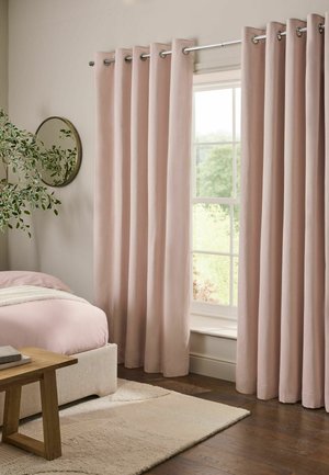 Weiche rosa Vorhänge hängen an einer silbernen Stange neben einem Fenster in einem Schlafzimmer mit einem beigen Bett, einer Holzbank, einem runden Spiegel und einer grünen Blattpflanze.