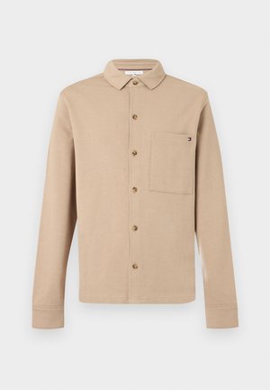 Beige langermet knappeskjorte med krage og en enkelt brystlomme med en liten logo på høyre side.