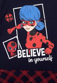T-shirt blu navy con un personaggio dei cartoni animati dai capelli blu e un outfit rosso con macchie nere, affiancato da un personaggio rosso. Il testo recita "CREDI in te stesso."