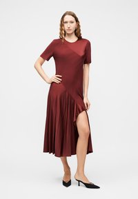 Γυναίκα με σγουρά μαλλιά που φοράει midi φόρεμα βουργουνδί με κοντά μανίκια, ασύμμετρο στρίφωμα και ψηλό άνοιγμα, συνδυασμένο με μαύρα mules με τακούνι.