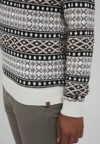Pull en tricot avec un motif géométrique en crème, marron et noir. Manches et ourlet côtelés, avec une petite étiquette en cuir près de l'ourlet.
