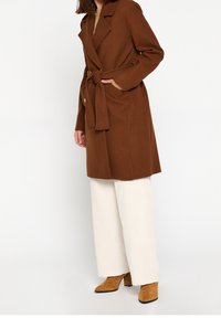 Vrouw draagt een bruine wollen jas met ceintuur, witte wijde broekspijpen en beige suède enkellaarsjes met hak tegen een effen witte achtergrond.