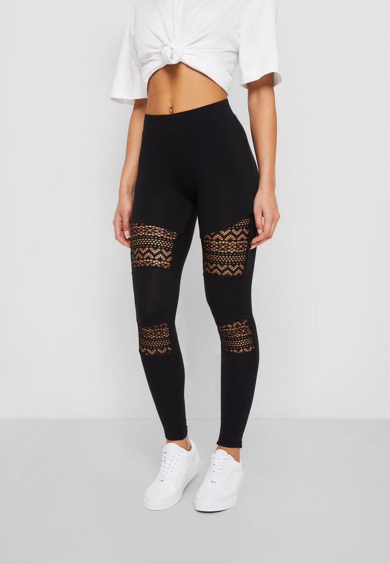 Leggings noirs avec des découpes en dentelle au niveau des genoux, présentant une texture lisse et un design taille haute, associés à des baskets blanches.