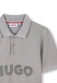 Grijze polo gemaakt van gestructureerde stof, met een kraag, drie knopen en een groot "HUGO" logo in donkerder grijs aan de voorkant.
