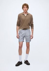 Jeune homme debout portant un pull marron à manches longues, un short bleu clair, des chaussettes blanches et des chaussures en cuir marron foncé sur un fond blanc uni.