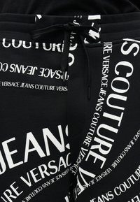 Μαύρο ύφασμα με επαναλαμβανόμενο λευκό κείμενο "Versace Jeans Couture" σε διάφορα μεγέθη και προσανατολισμούς, με μαύρο κορδόνι δεμένο στη μέση.