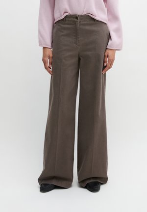 Personne portant un pantalon large marron, un haut à manches longues rose clair et des chaussures noires, debout devant un fond uni.