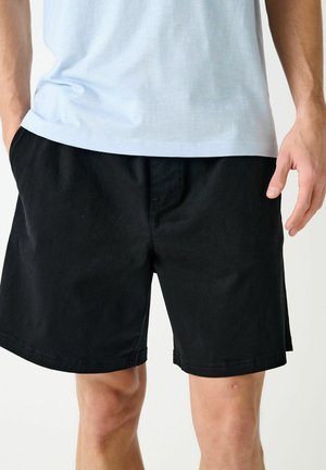 Mies, jolla on yksiväriset mustat polvipituiset shortsit ja vaaleansininen t-paita, kädet taskuissa.