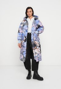 Manteau matelassé floral avec un mélange de tons bleus et roses, capuche, fermeture éclair, poignets côtelés et grandes poches, porté par un mannequin portant un pantalon sombre.
