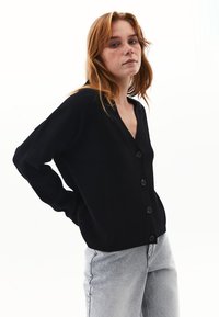 Musta neuletakki, joka on valmistettu pehmeästä neulekankaasta, sisältää V-pääkohdan, etupainikkeet ja löysän istuvuuden, jossa on ribbaatut hihansuut. Yhdistetty vaaleanharmaisiin farkkuihin.